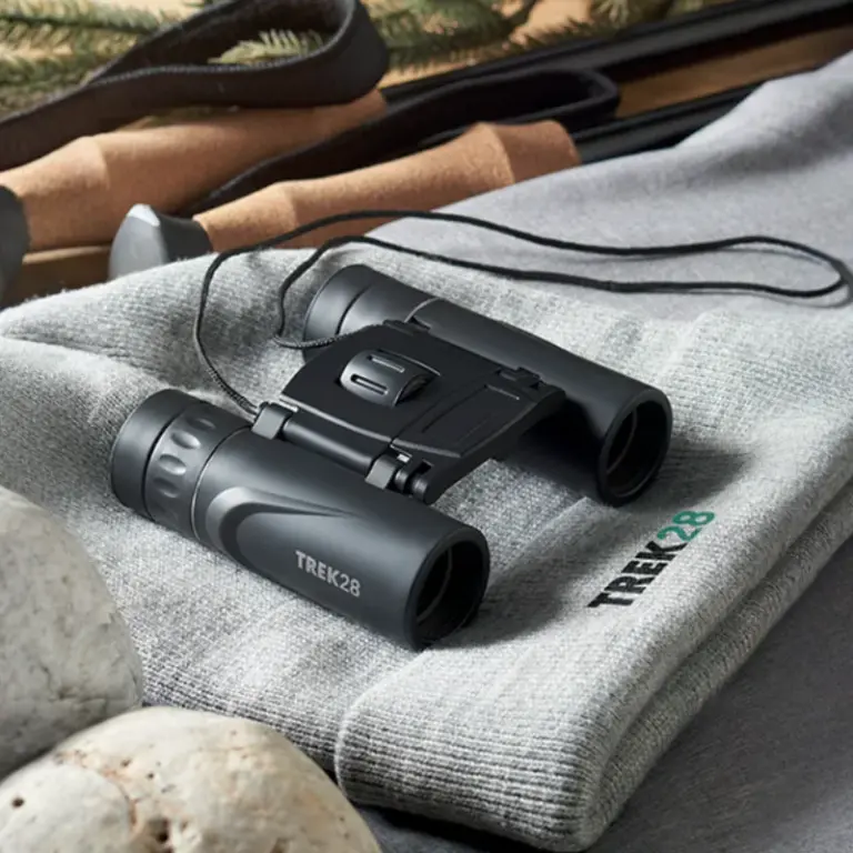 Binoculares Trek28 sobre gorro gris, con bastones de senderismo y piedras.