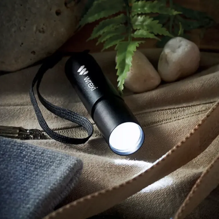 Una linterna negra "Wrexi" ilumina un entorno con piedras, una hoja y tela.