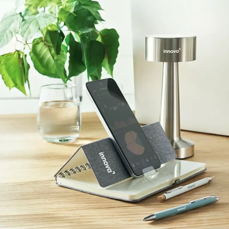 Un escritorio ordenado con un móvil apoyado en una libreta, bolígrafos, lámpara y planta.