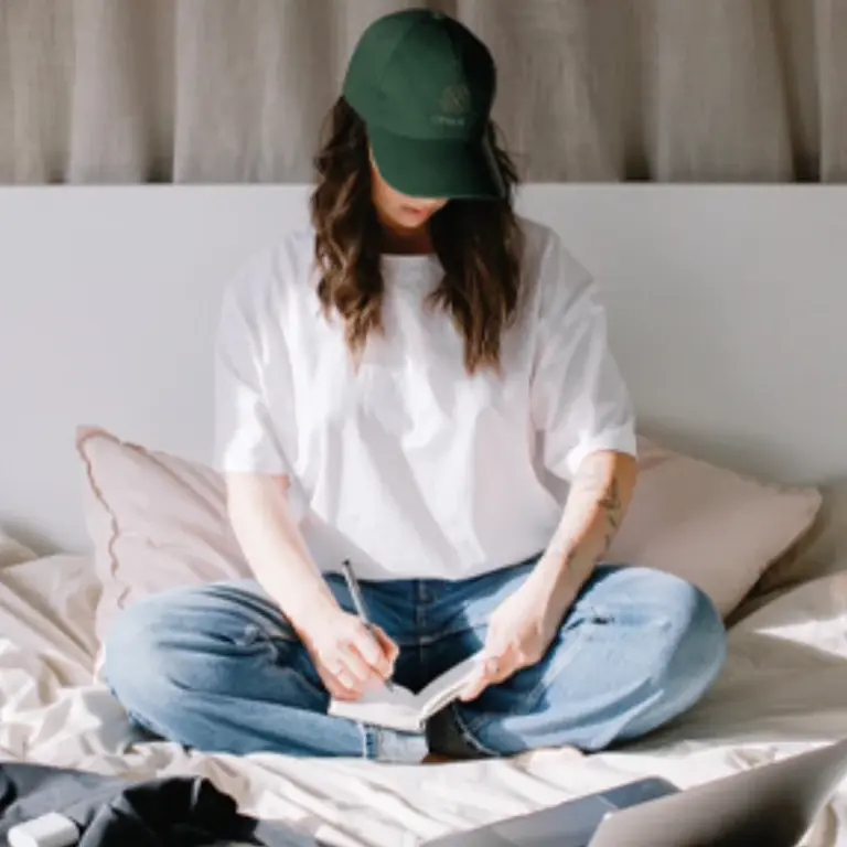 Mujer con gorra verde escribe en un cuaderno sentada en la cama con un portátil cerca.