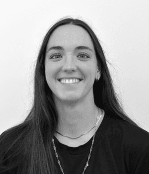 La imagen es un retrato en blanco y negro de una joven sonriendo. Tiene el pelo largo y oscuro, y lleva una camiseta oscura y collares. El fondo es blanco.