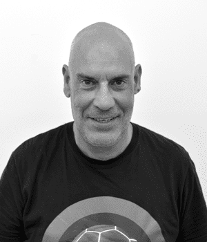 La imagen muestra un retrato en blanco y negro de un hombre calvo con una camiseta oscura que tiene un diseño circular en el pecho. El hombre mira directamente a la cámara y parece sonreír ligeramente. El fondo es blanco y liso.