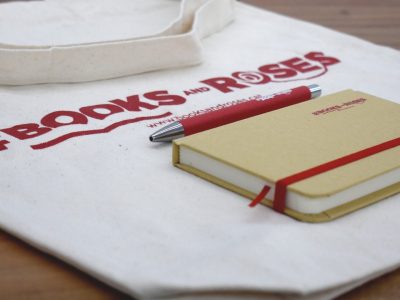 Bolsa de tela con logo, libreta y bolígrafo rojo sobre una mesa de madera.